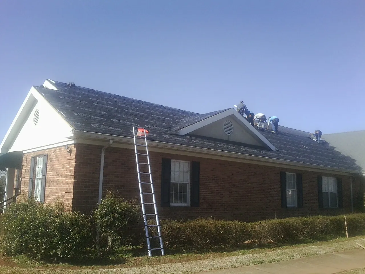 Crew tearing shingles off a brick ranch roof with a ladder set — Emergency Roof Repair work by our crew in Cashion
