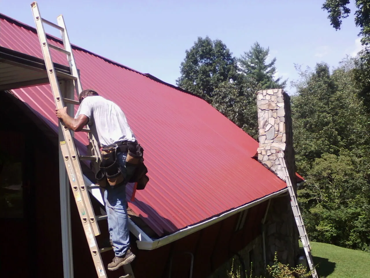 Roofer on a ladder beside a red metal roof and stone chimney — Soffit & Fascia Repair by Roofing Brothers in Cashion